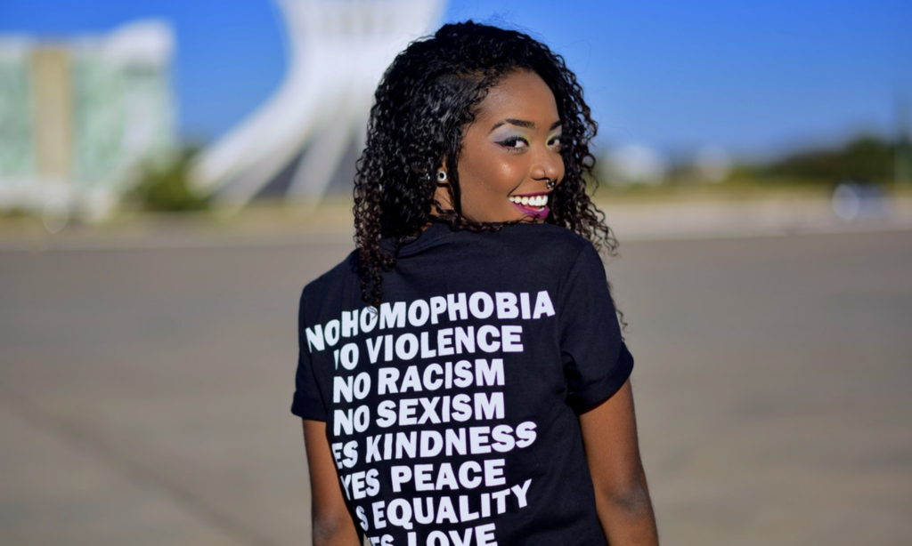 A black women posing with her back to us wearing a black shirt with white writing that says no homophobia, no violence, no racism, no sexism, yes kindness, yes peace, yes equality, yes love