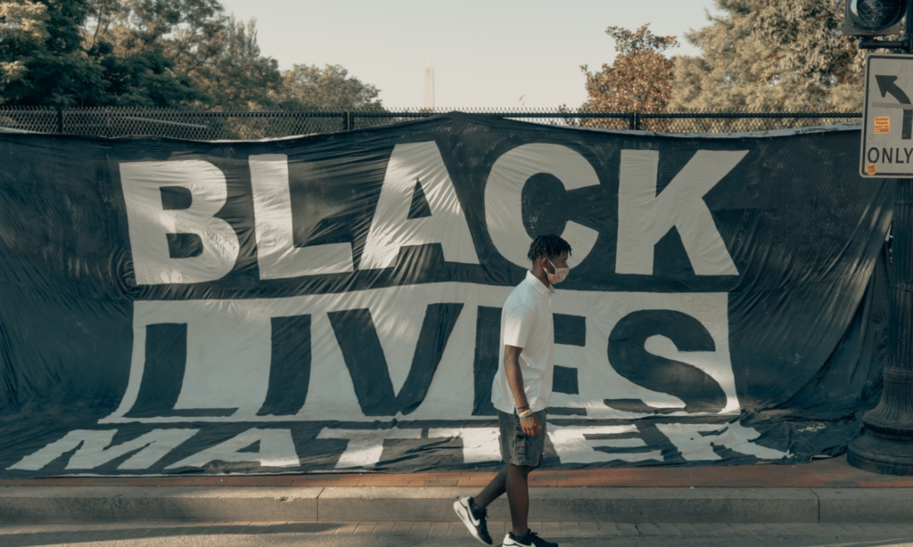 Black man walking on the street with a black lives matter banner on the sidewalk